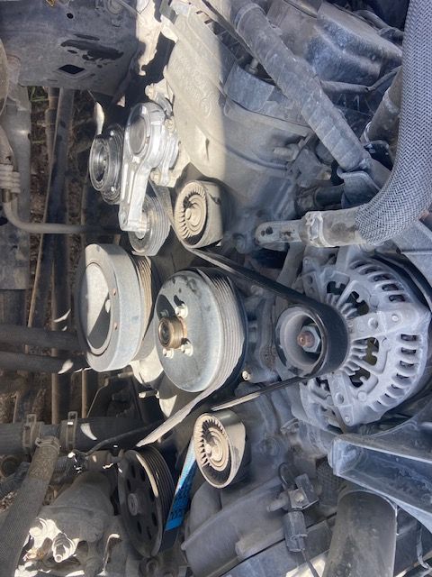 Close-up of a car engine's pulleys and belts. The engine is dirty and has various components visible.