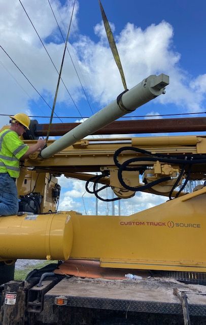 Construction worker in yellow vest installing a long, cylindrical drill bit on a yellow machine, near power lines.