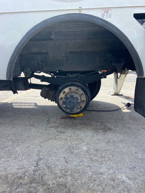 Underside of a white truck showing the rear axle, wheels, suspension, and leaf springs on a concrete surface.