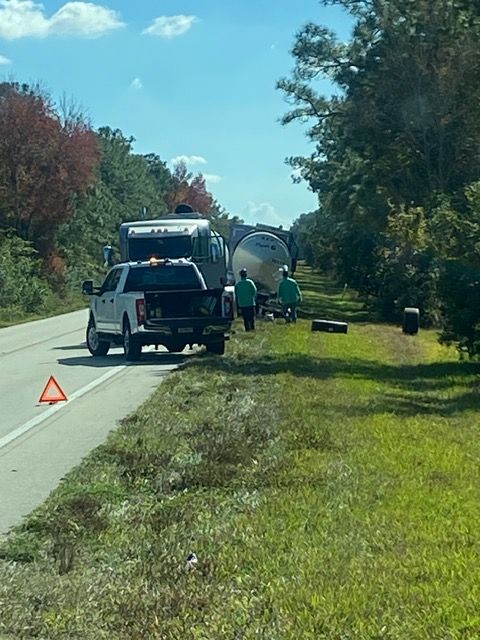 A tanker truck and pickup truck are on the side of a road. People in green shirts are near the tanker. Orange hazard cone.