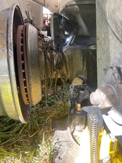 Close-up of a damaged truck wheel with a brake rotor, wires, and debris, set in grass.