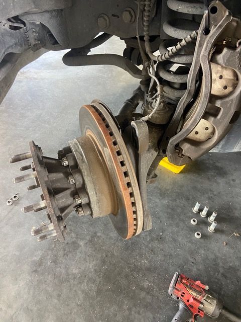 A vehicle's front wheel assembly disassembled; brake rotor, caliper, and hub exposed. Tools and bolts visible.