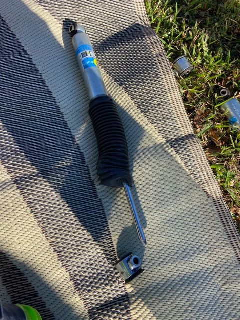 A silver and blue Bilstein shock absorber with a black protective boot, lying on a striped mat outdoors.