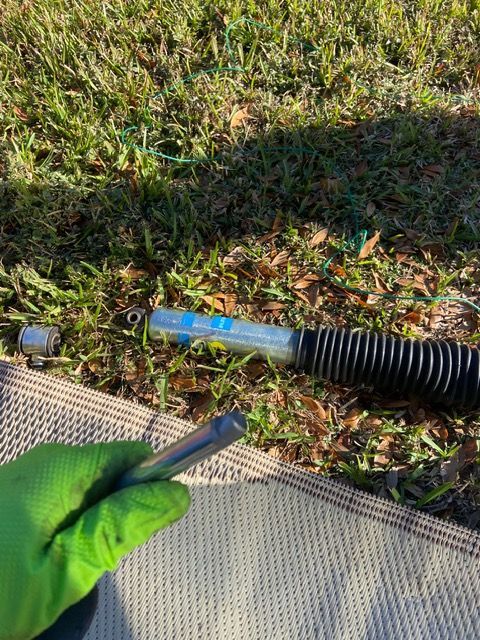 Green-gloved hand holds a metal tool near a blue and silver shock absorber lying on grass with a protective cover.