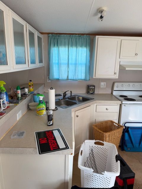 A cluttered kitchen with white cabinets, light-colored countertops, a window with blue curtains, and a white stove.