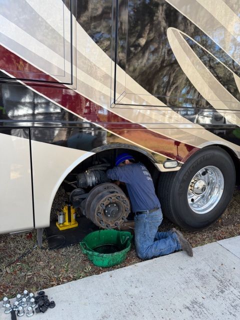 Mechanic working on the wheel of a large RV; he's kneeling, a green pan beneath, and tools nearby.