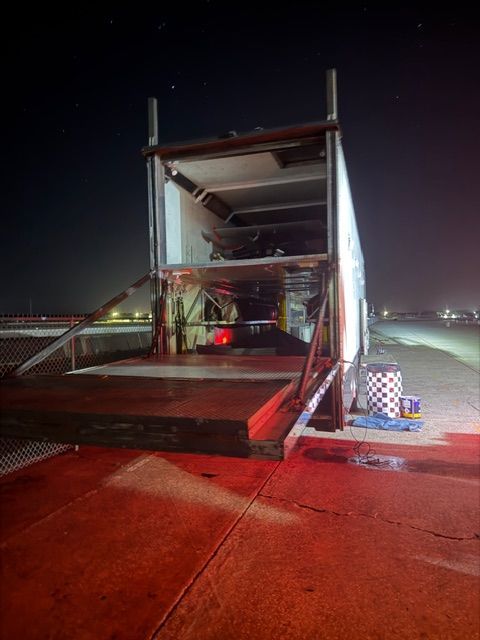 A race car trailer with its lift gate lowered at night on a track, with equipment sitting on the asphalt nearby.