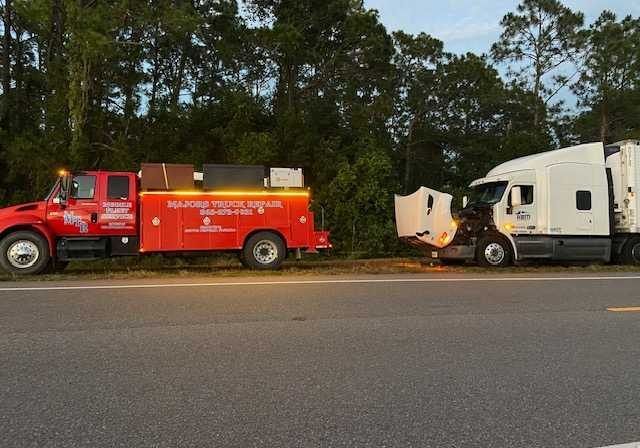 A red tow truck is parked next to a white semi truck on the side of the road.
