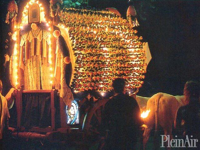 Processione notturna, con una struttura decorata illuminata da numerose candele e fiori. La gente osserva.