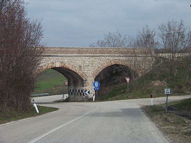 Ponte di pietra sopra una strada di campagna con aperture ad arco. Il cielo è coperto.