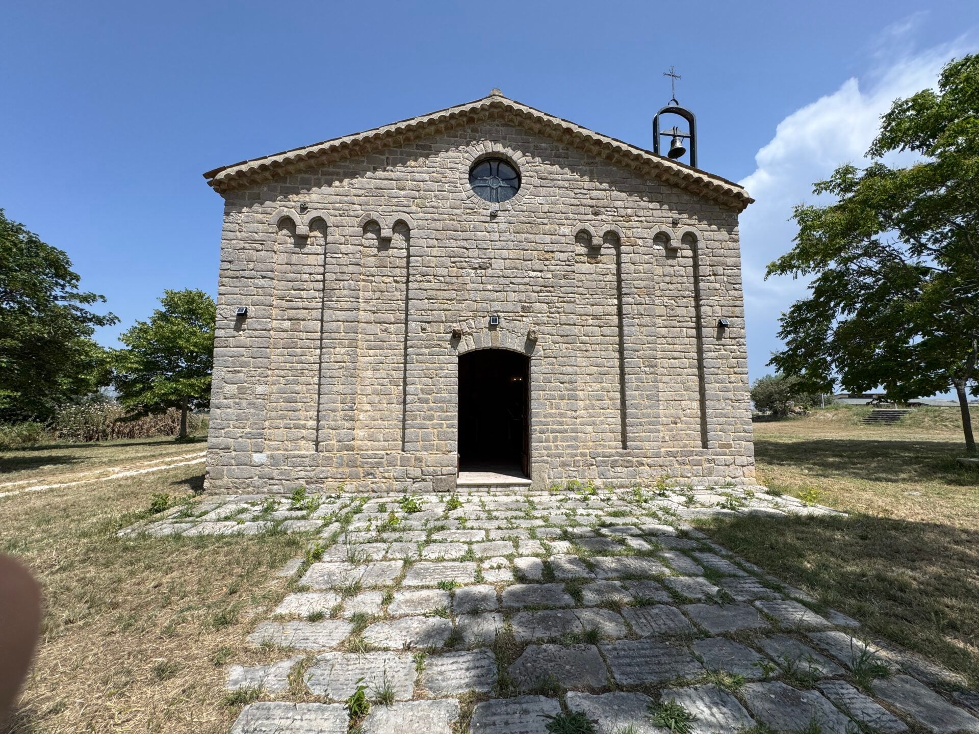 Chiesa in pietra con portale ad arco, finestra rotonda, piccolo campanile e sentiero in pietra. Cielo azzurro.