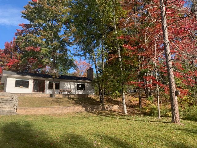 Cottage with a white exterior sits in front of trees with green and red fall foliage.