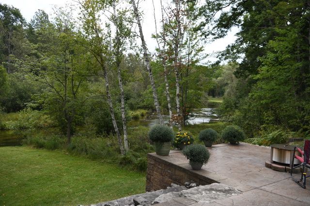 A scenic outdoor view. Green lawn and trees frame a river. Four potted plants sit on a patio.