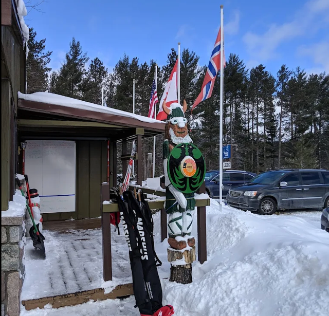 Entrance to a ski lodge with flags, a wooden statue, and snow-covered ground. Skis lean against the building.