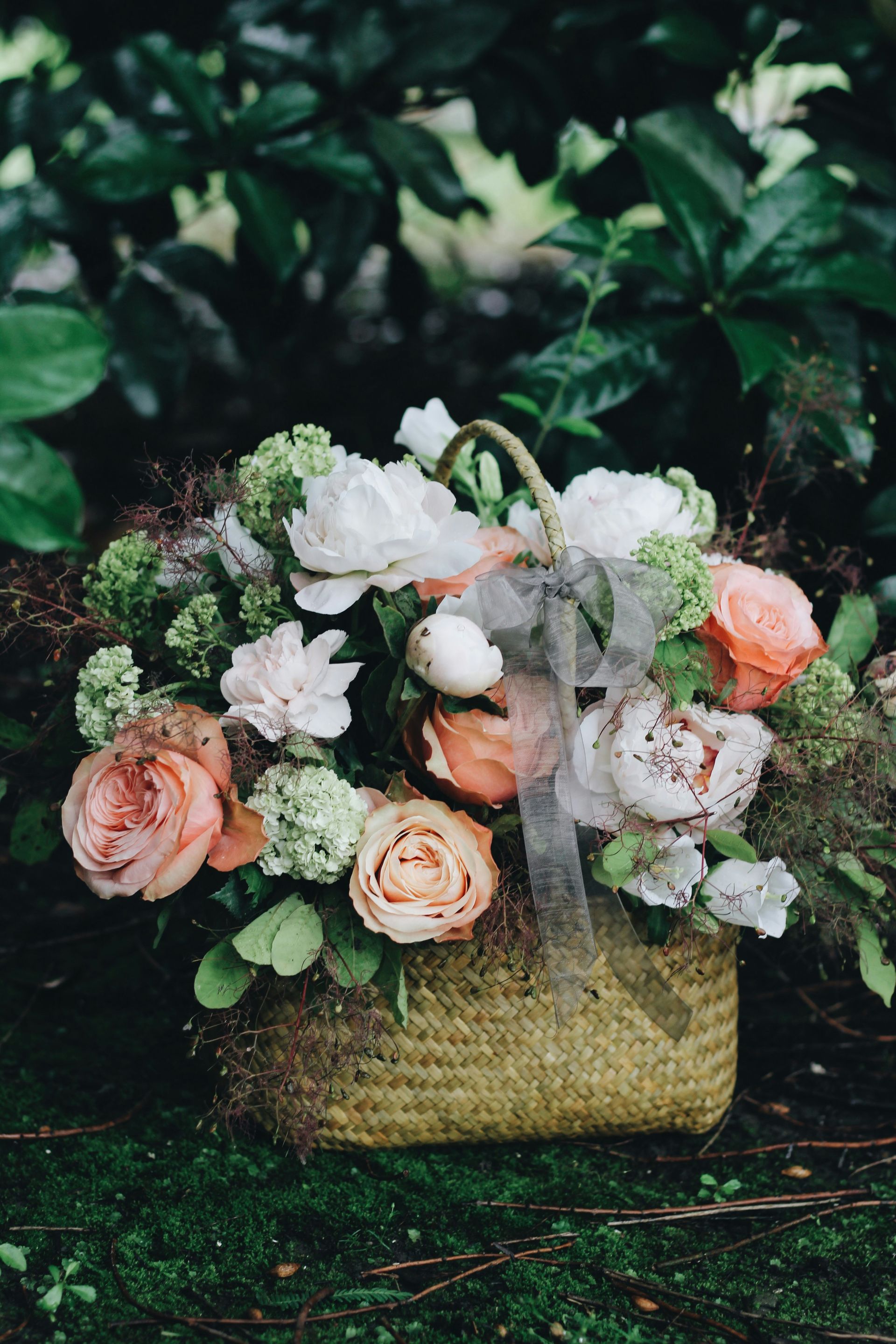 Basket of peach and white flowers with greenery, set on a mossy surface.