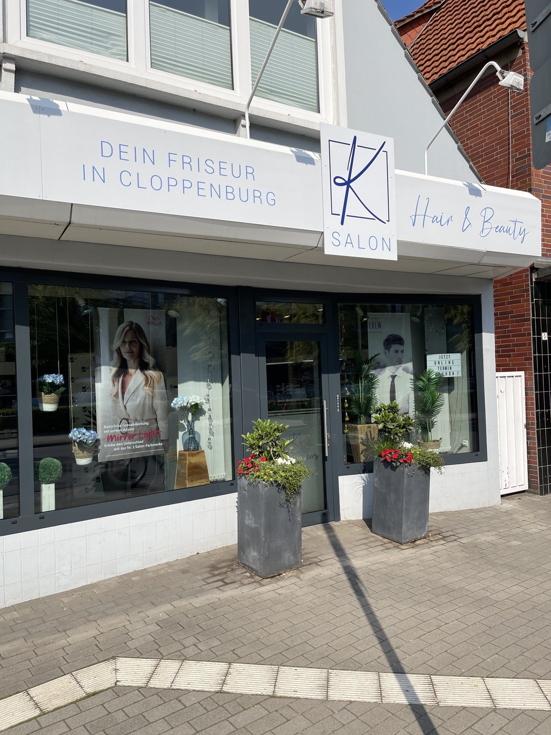 Ein Friseursalon, „De Knipschuur“, vor einem Gebäude mit grauer Fassade, Blumentöpfen und Bürgersteig.