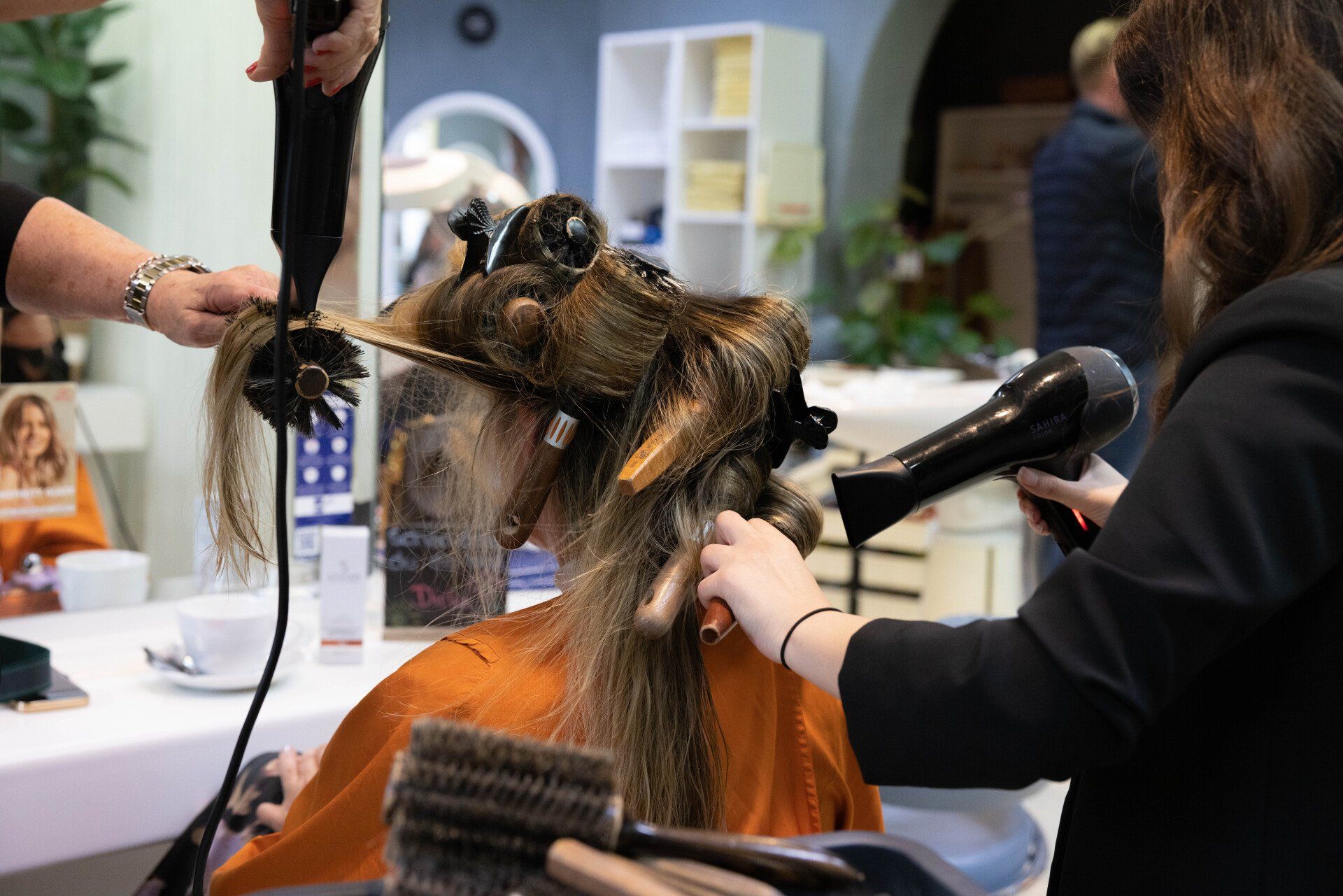 Zwei Stylisten föhnen in einem Salon das lange, hellbraune Haar einer Kundin.