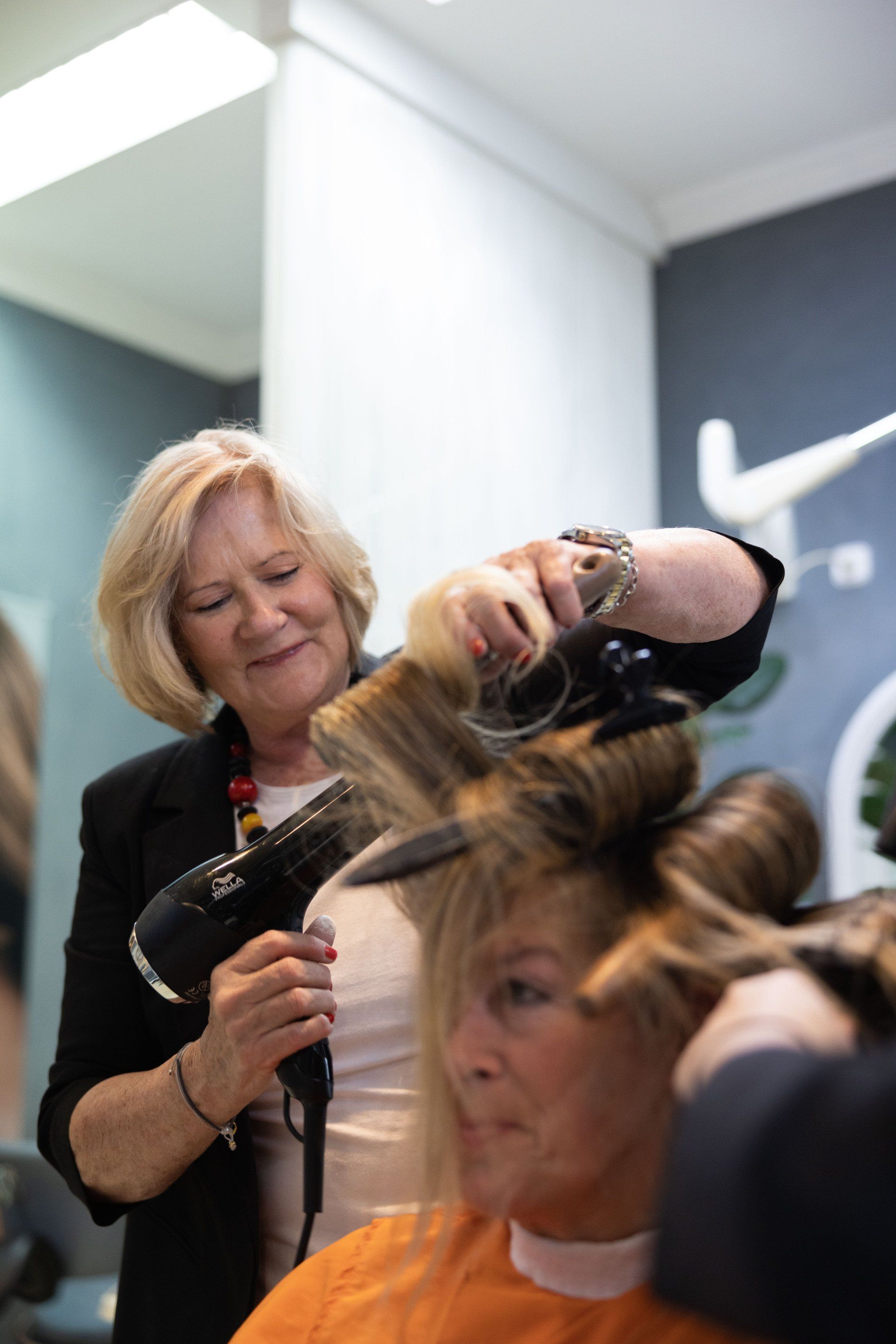 Eine Friseurin föhnt einer Kundin in einem Salon mit einem schwarzen Haartrockner die Haare. Beide Frauen sind blond.
