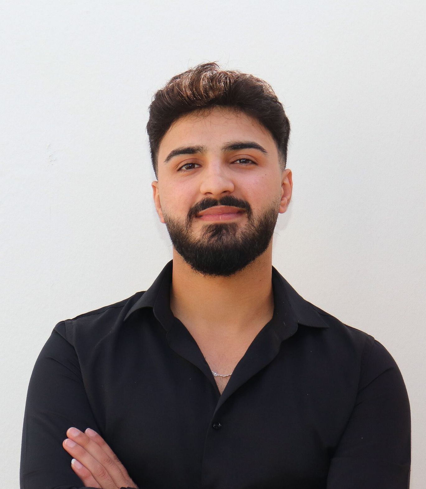 Mann mit dunklem Haar und Bart, trägt ein schwarzes Hemd, die Arme sind verschränkt und er lächelt vor einer weißen Wand.