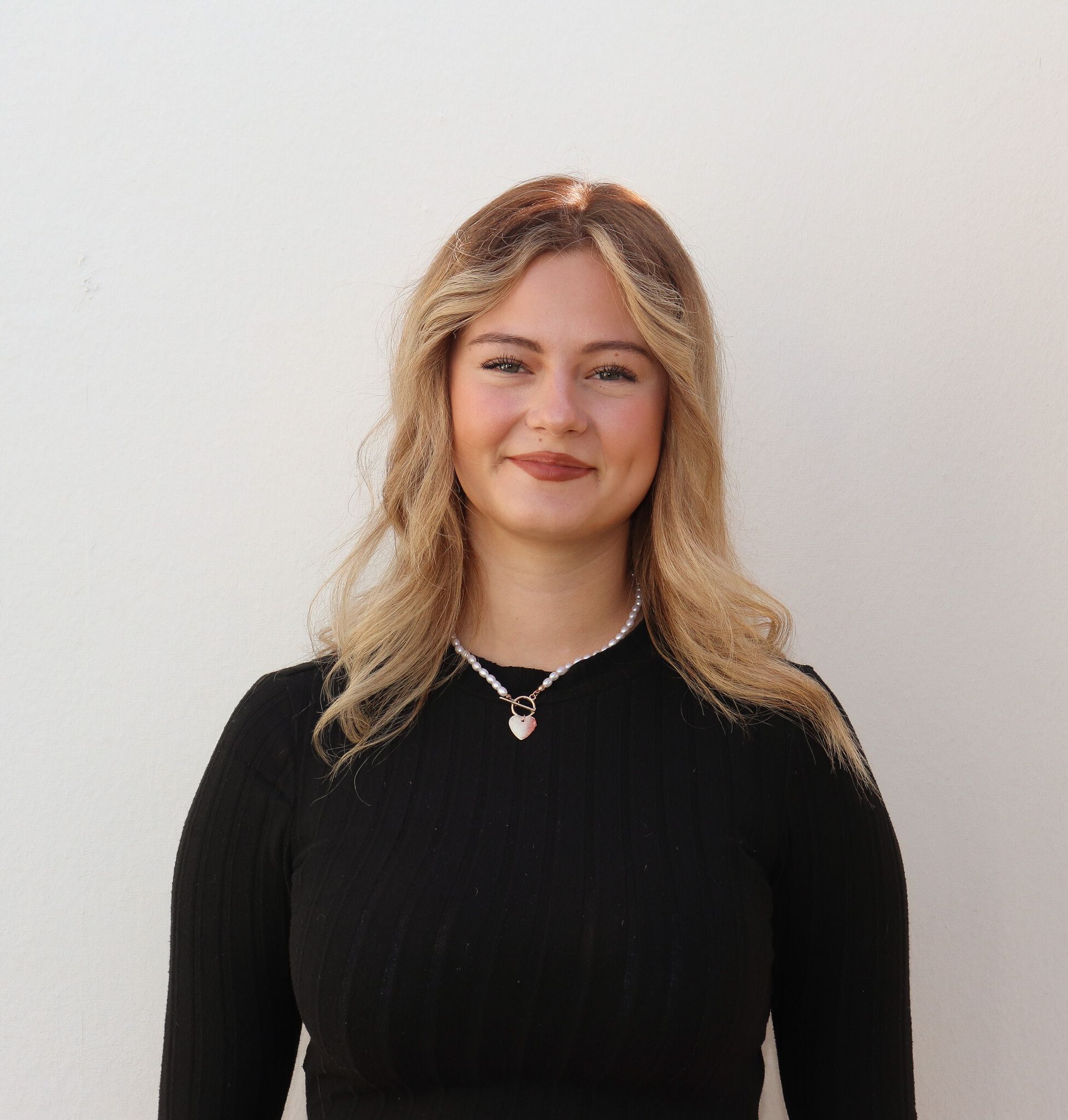 Lächelnde Frau mit blondem Haar, schwarzem Oberteil und Halskette, steht vor einer weißen Wand.