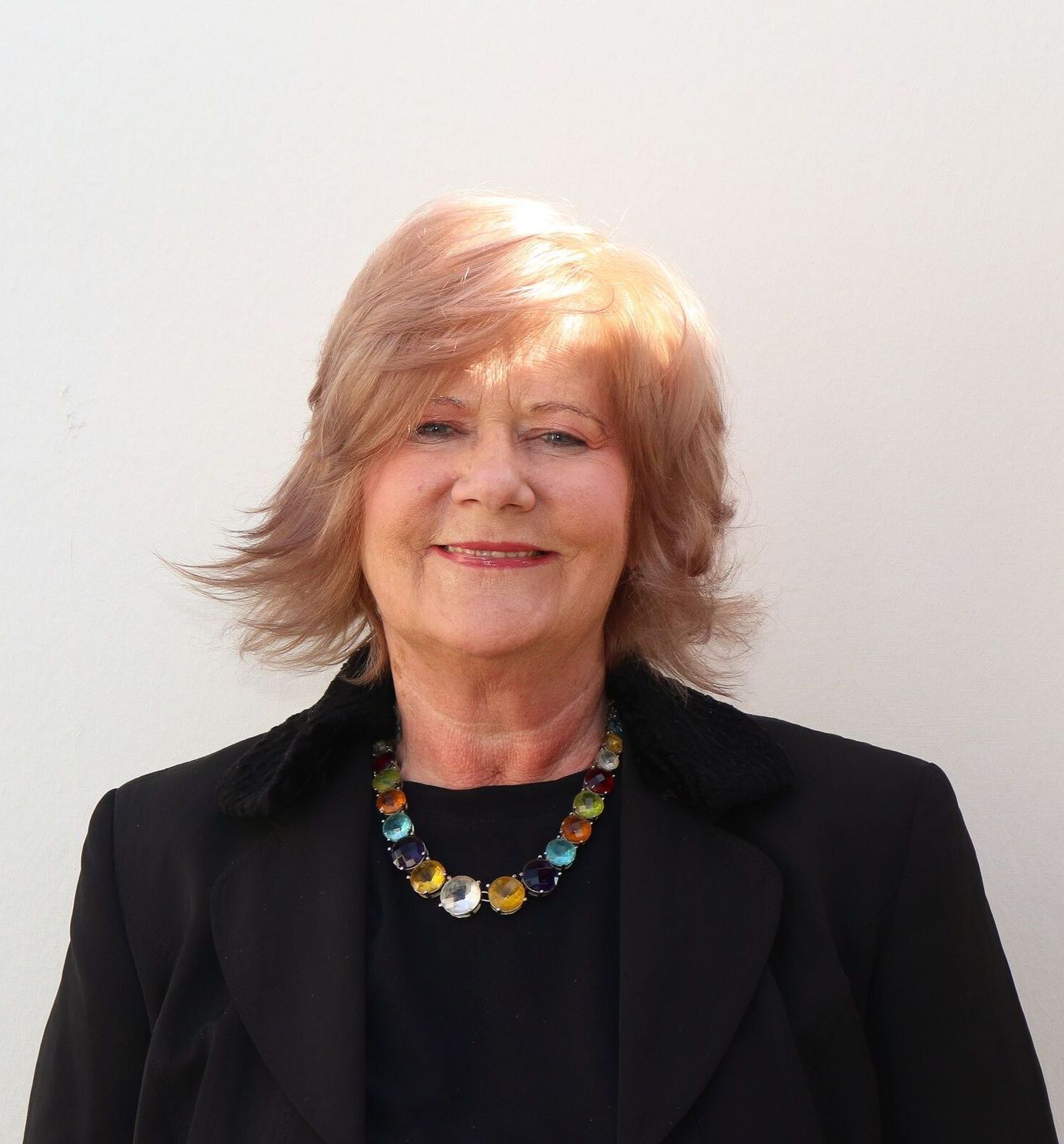 Frau mit blonden Haaren und bunter Halskette, lächelnd vor einer weißen Wand.