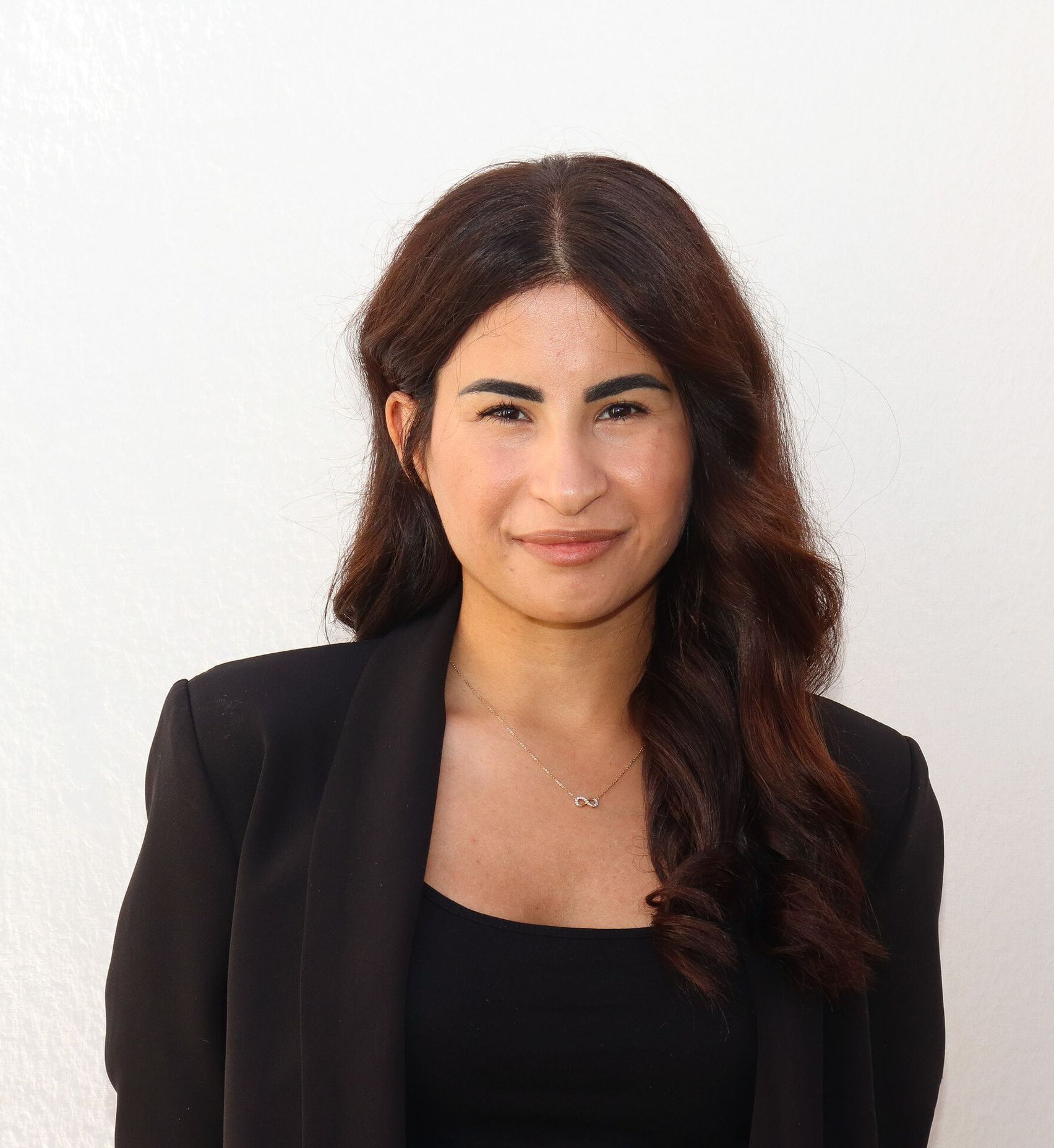 Frau mit dunklem Haar, trägt einen schwarzen Blazer und ein Top und lächelt in die Kamera. Vor weißem Hintergrund.