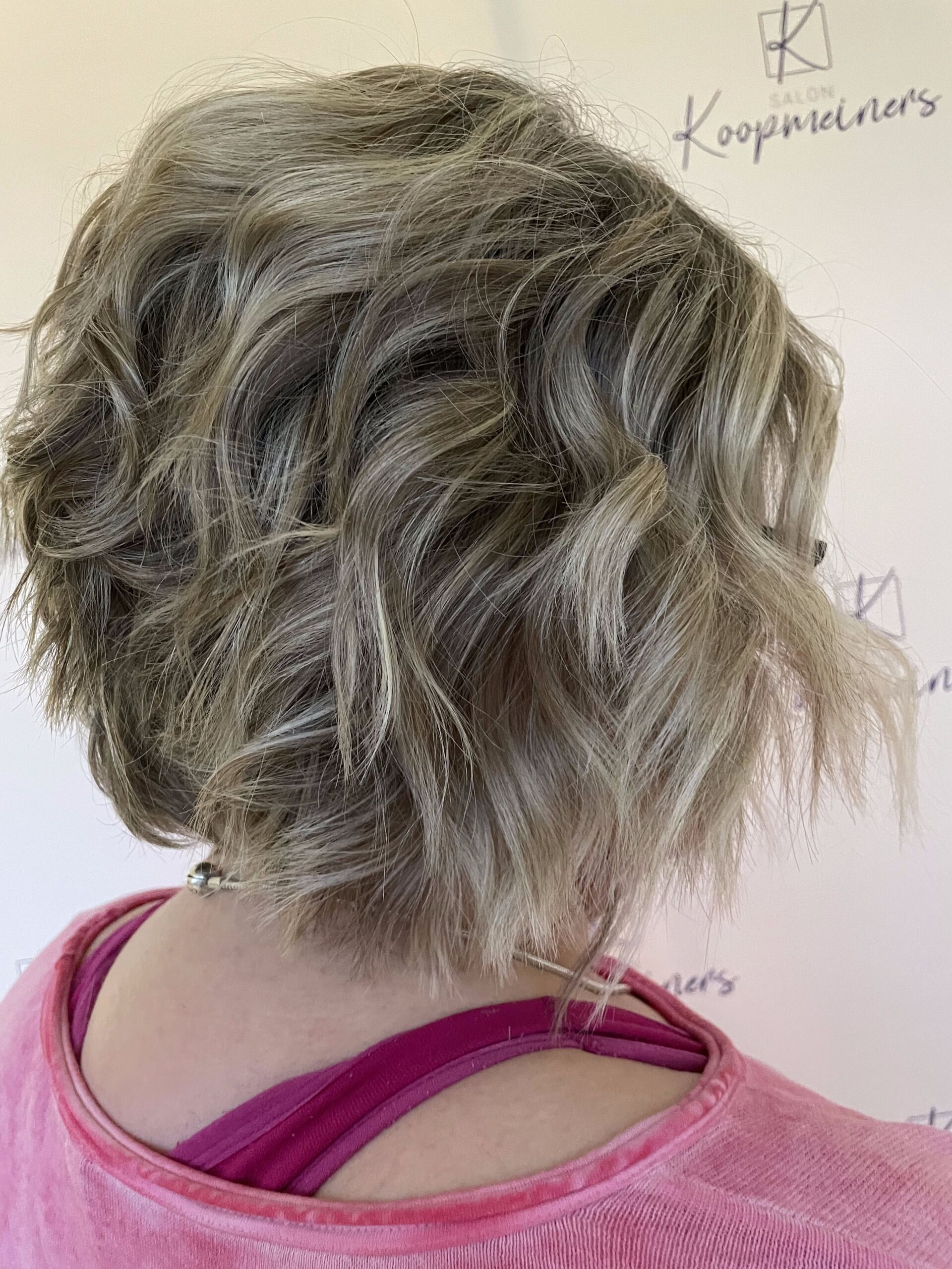 Frau mit kurzen, welligen, grauen und blonden Haaren, trägt ein rosa Hemd.