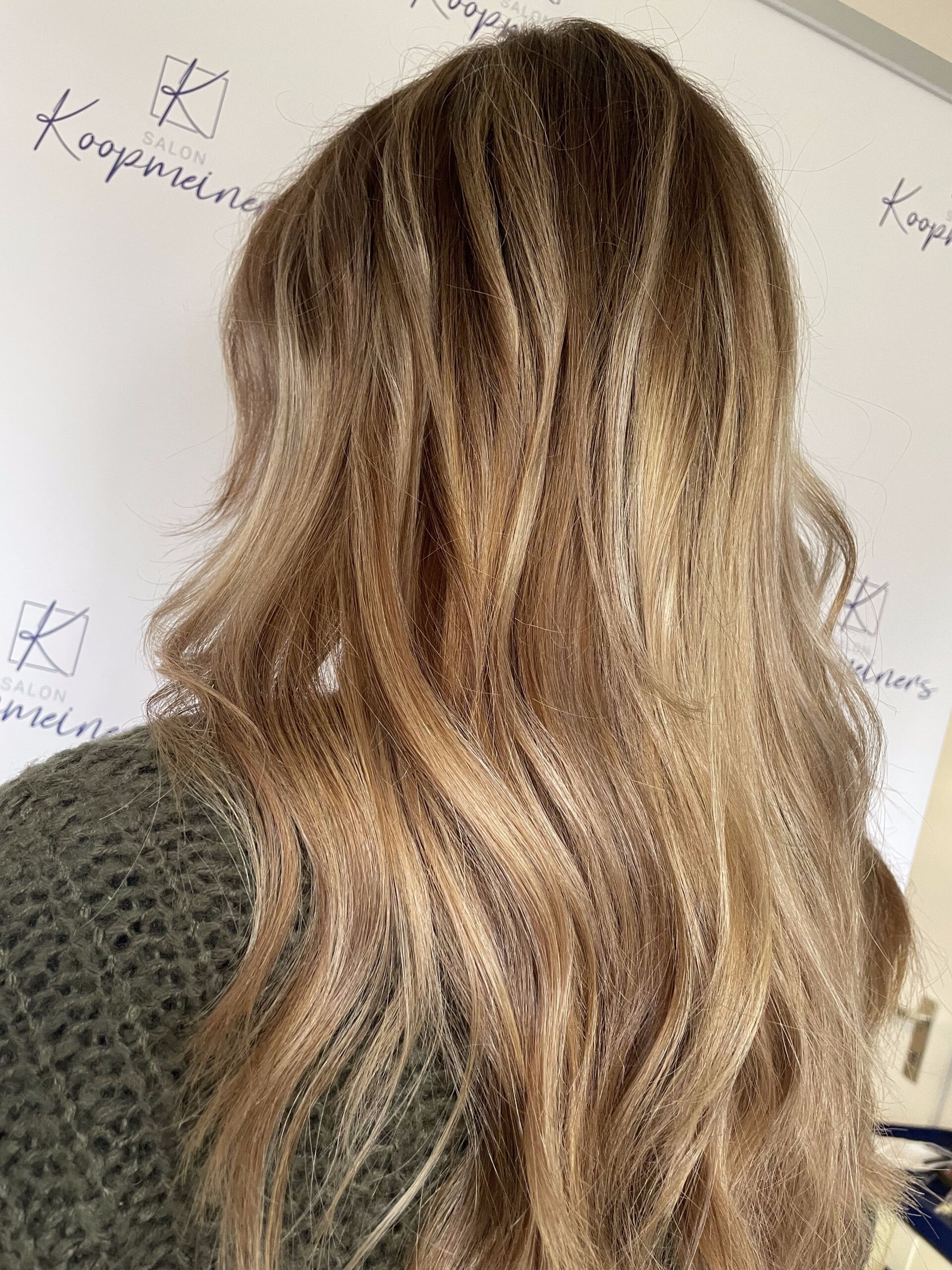 Frau mit welligem, blondem Haar, gesträhnt, trägt einen grünen Pullover und steht im Haus.