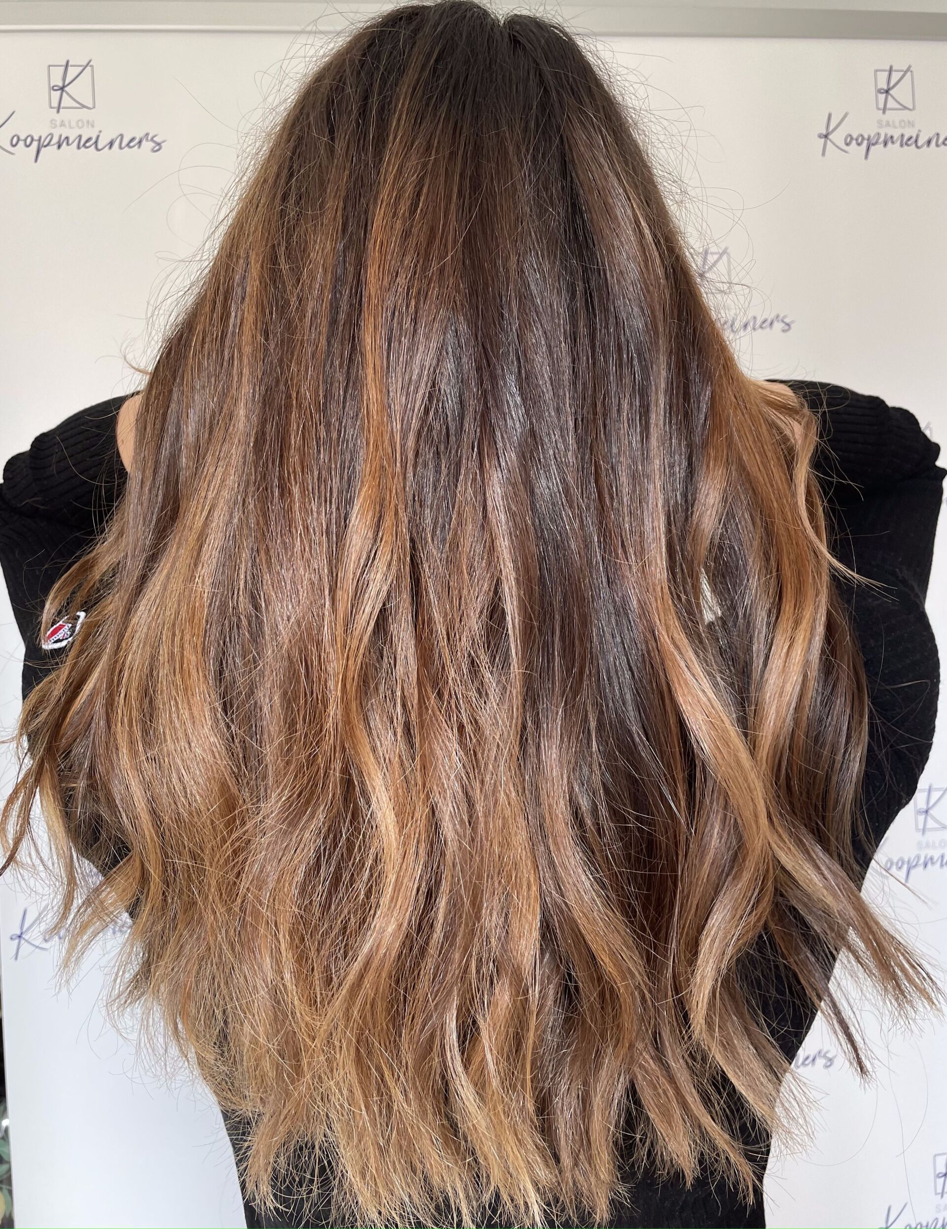 Frau mit langem, welligem braunem Haar, helleren Strähnchen, trägt einen schwarzen Pullover.