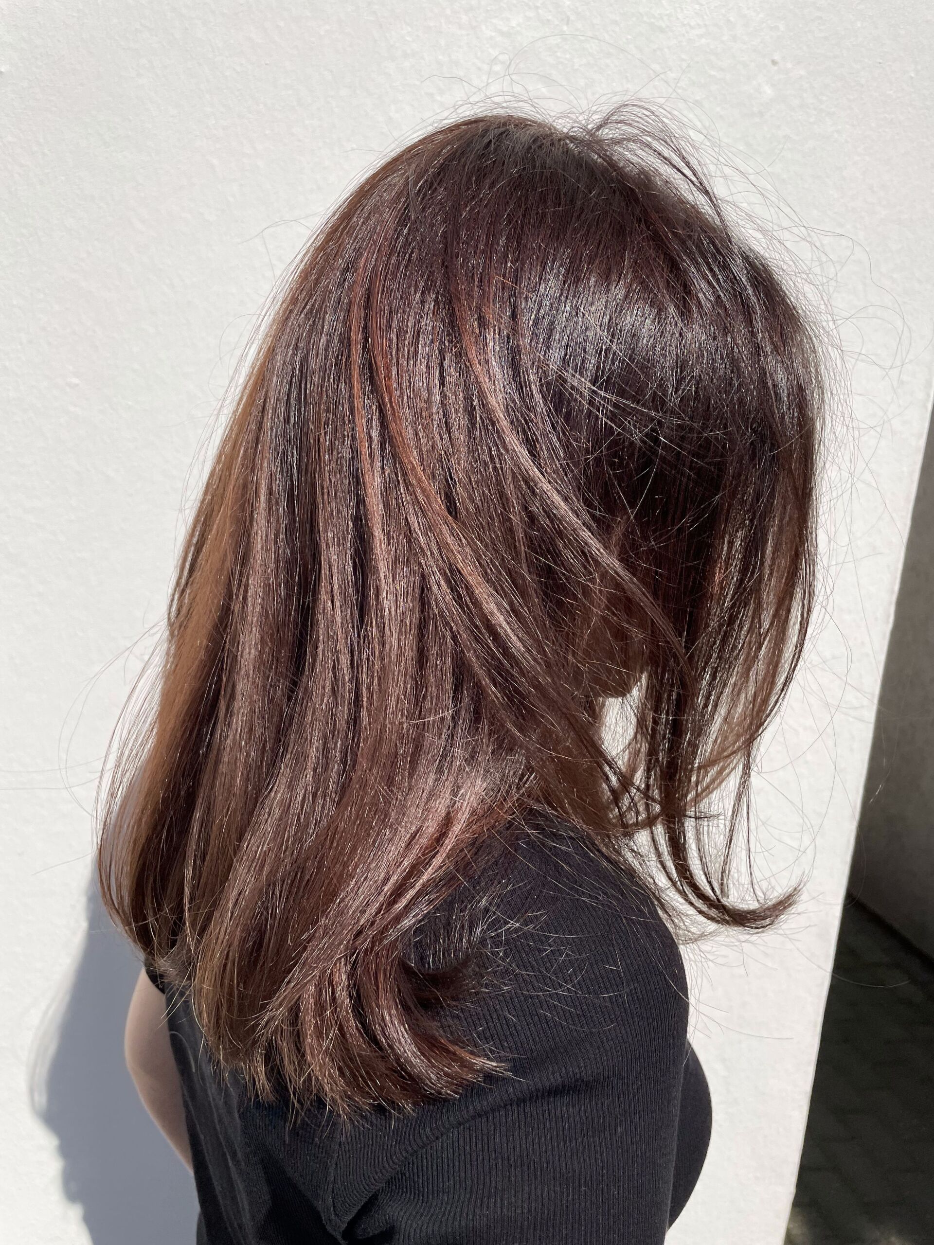 Frau mit braunem Haar, Sonnenlicht, trägt ein schwarzes Oberteil, vor einer weißen Wand.