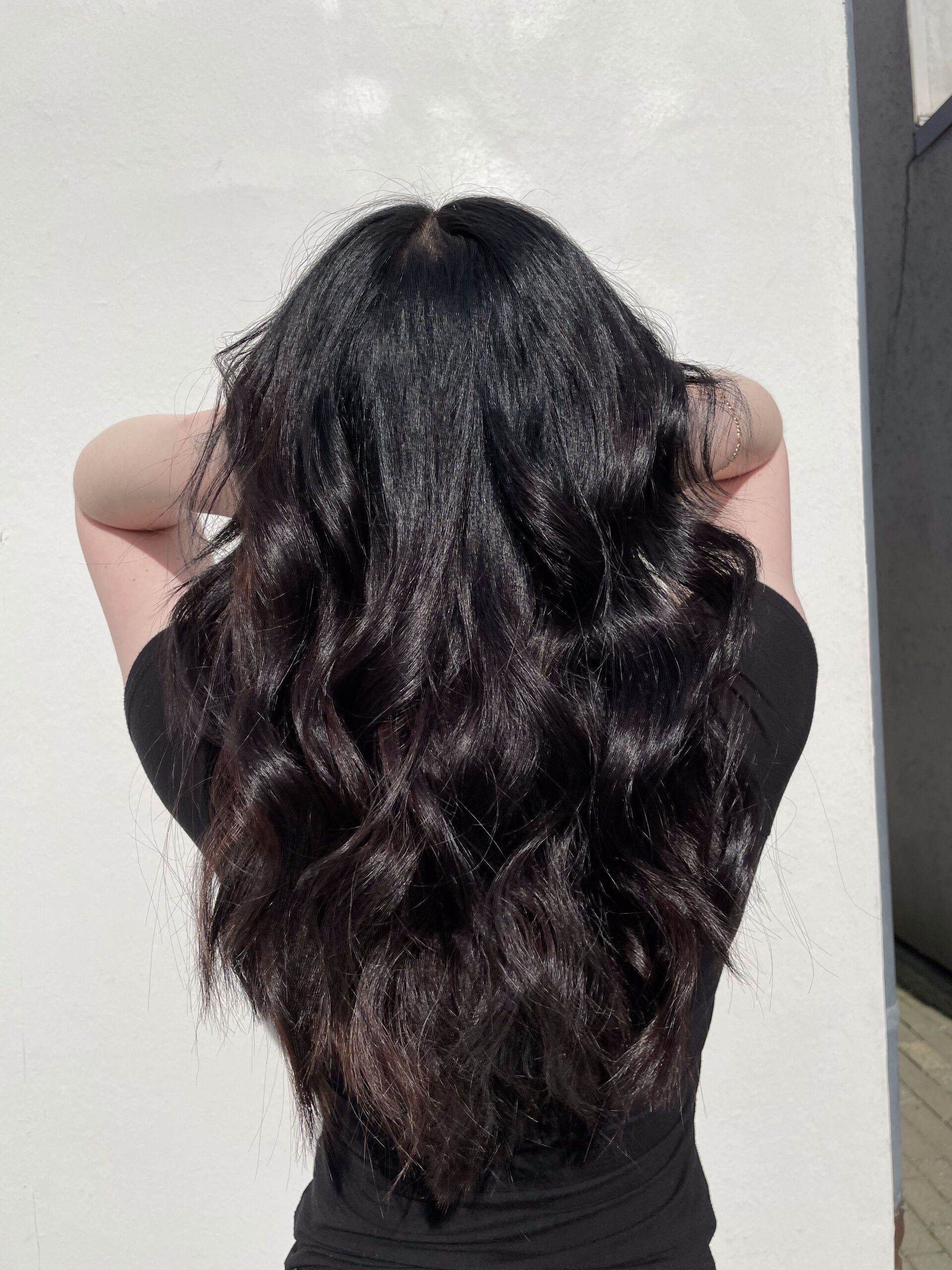 Frau mit langem, dunklem, welligem Haar, erhobenen Armen, steht vor einer weißen Wand.