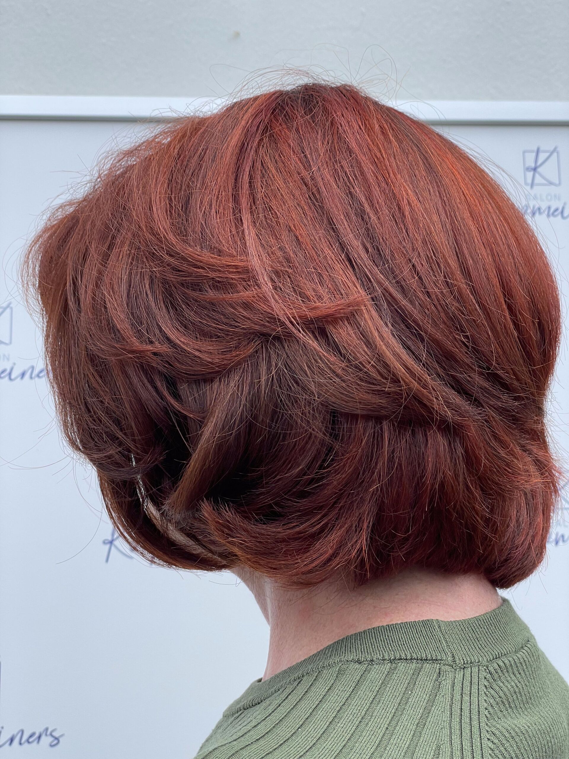 Frau mit gestuftem, rotbraunem Haar und grünem Pullover, von hinten betrachtet.