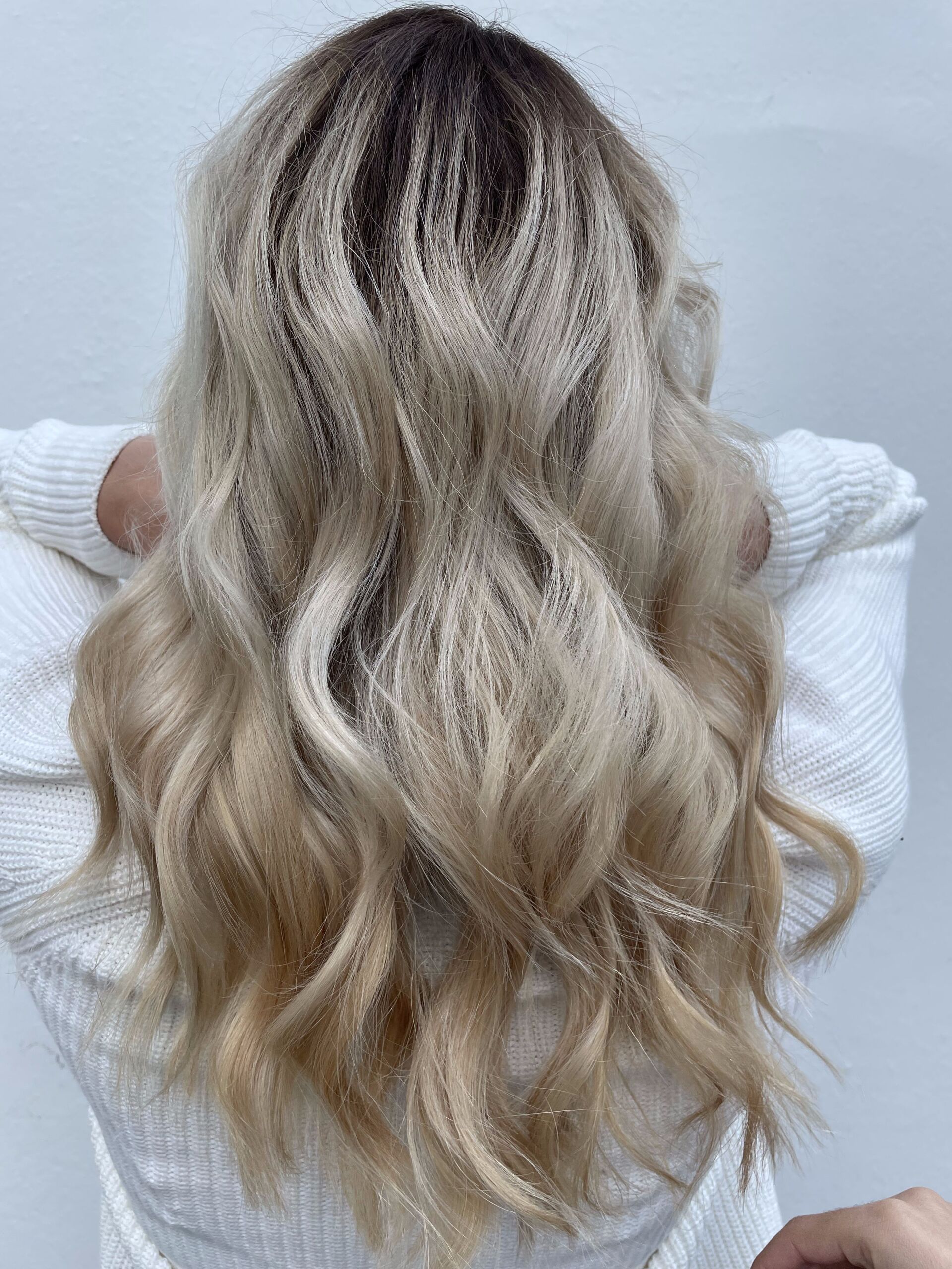 Frau mit langen, welligen blonden Haaren, dunklen Haarwurzeln, in einem weißen Pullover.