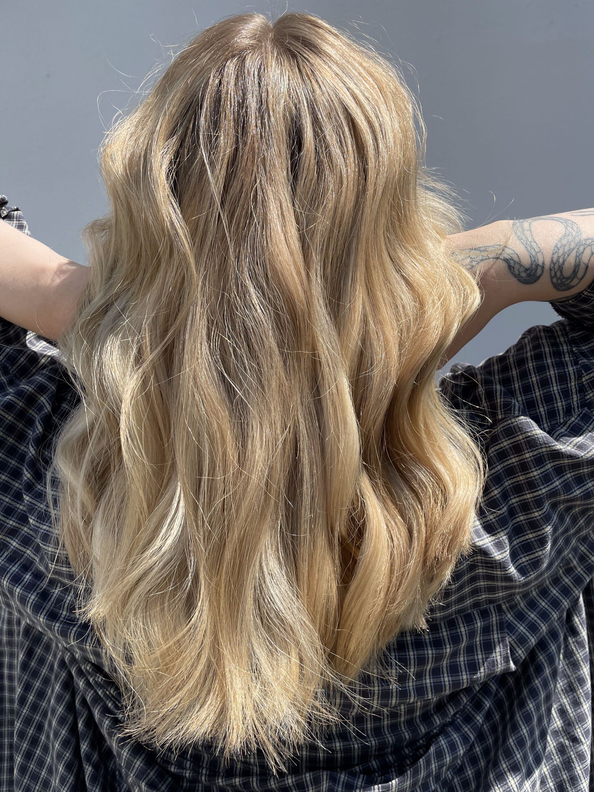 Blondes, welliges Haar, Rückansicht, Hände hinter dem Kopf, trägt ein kariertes Hemd.