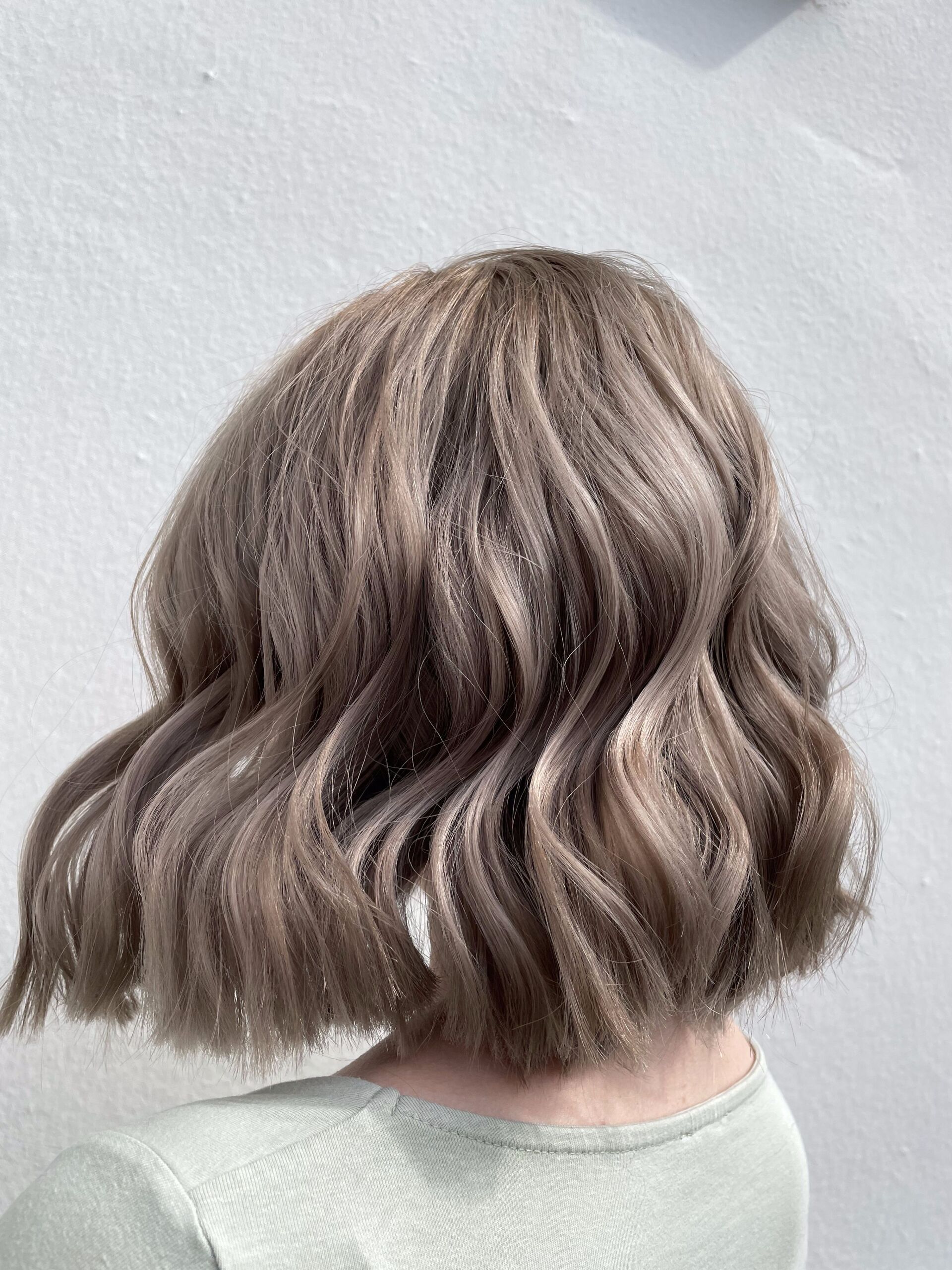 Schulterlanges, welliges, aschbraunes Haar. Rückansicht einer Person vor einer weißen Wand.