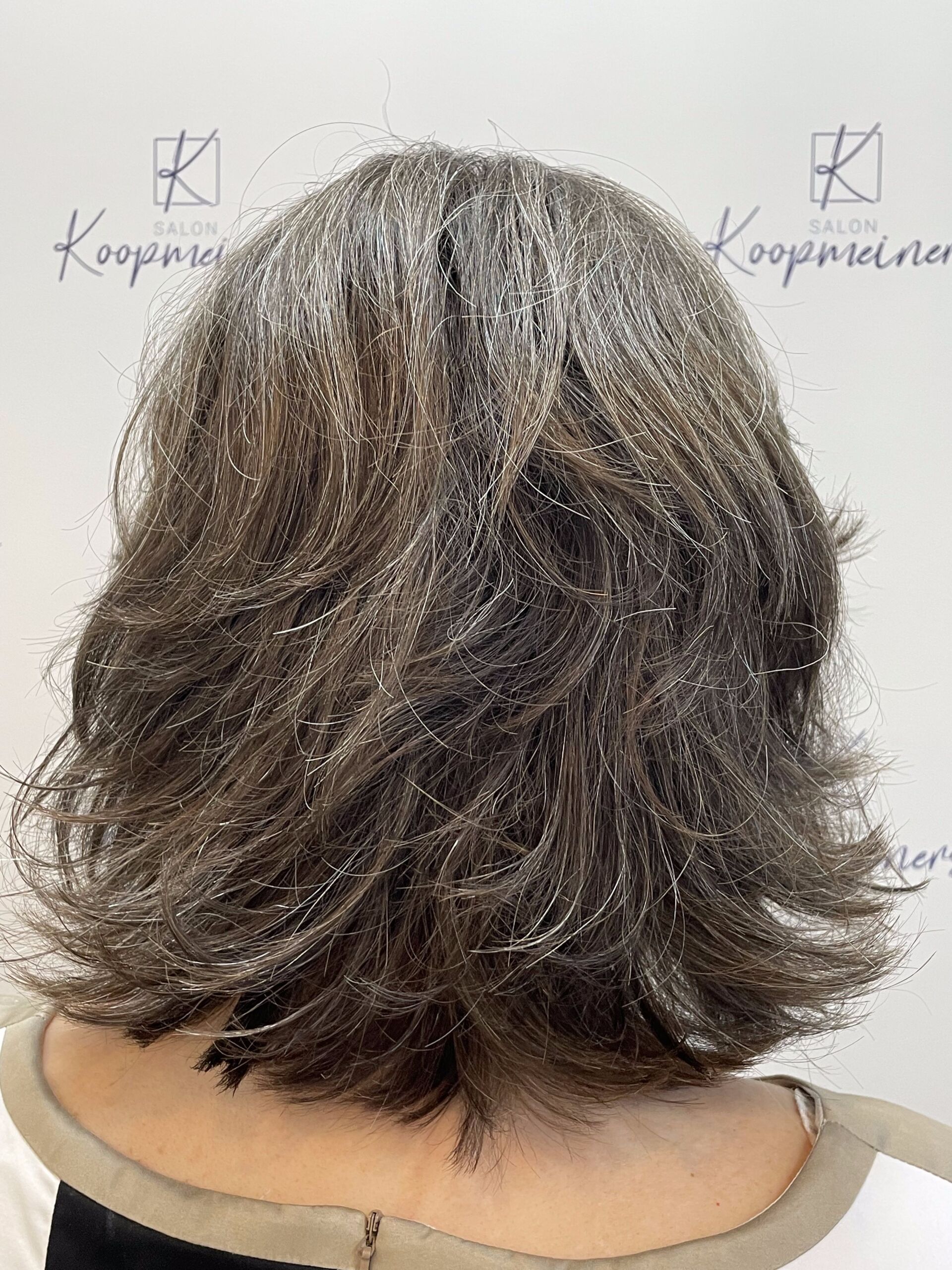Rückansicht einer Frau mit gestuftem, ergrauendem braunem Haar. Sie ist in einem Salon; im Hintergrund ist ein Logo zu sehen.