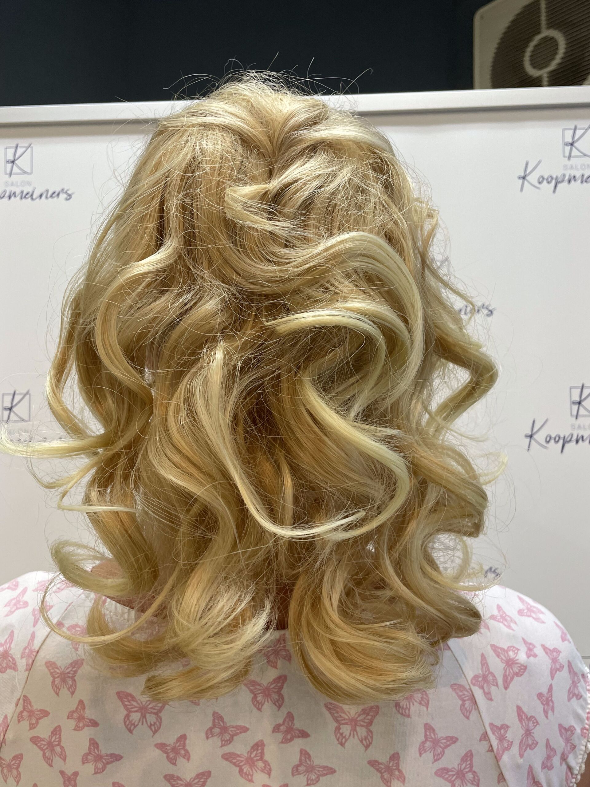 Blondes Haar in lockeren Wellen gestylt, mit einem weiß-rosa gemusterten Oberteil und weißem Hintergrund.