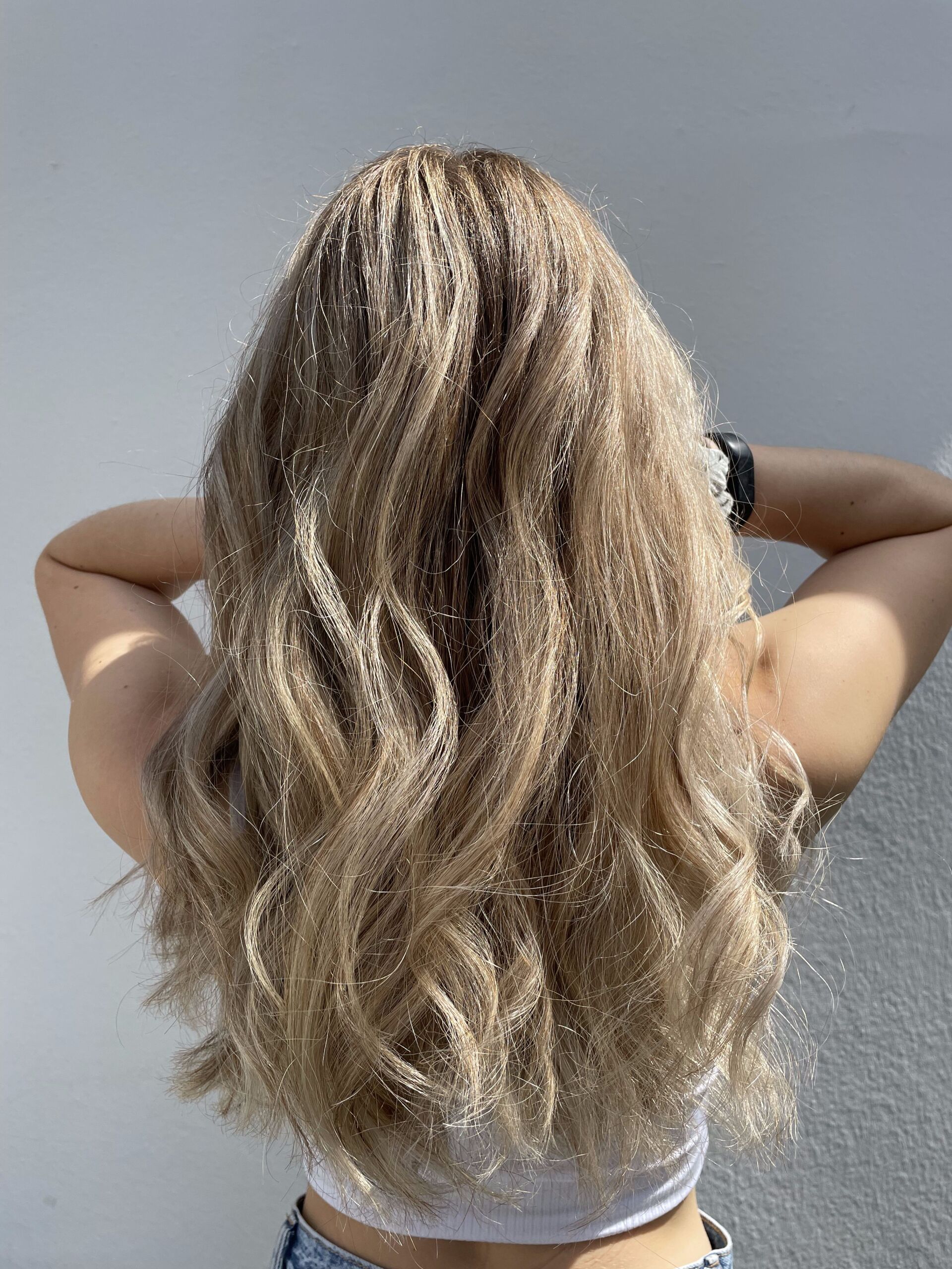 Frau mit langem, welligem blondem Haar, erhobene Arme, vor einer weißen Wand.