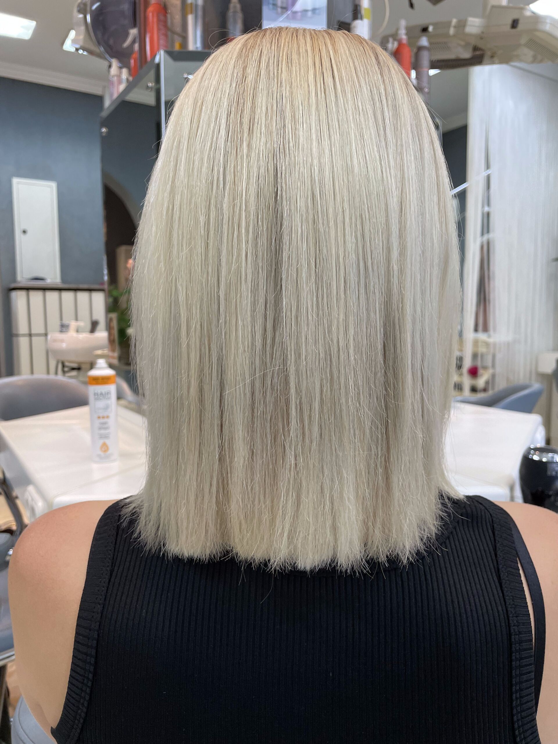 Blondes Haar mit sichtbaren Haarwurzeln, zu einem schulterlangen Bob frisiert. Frau trägt ein schwarzes Tanktop, drinnen.