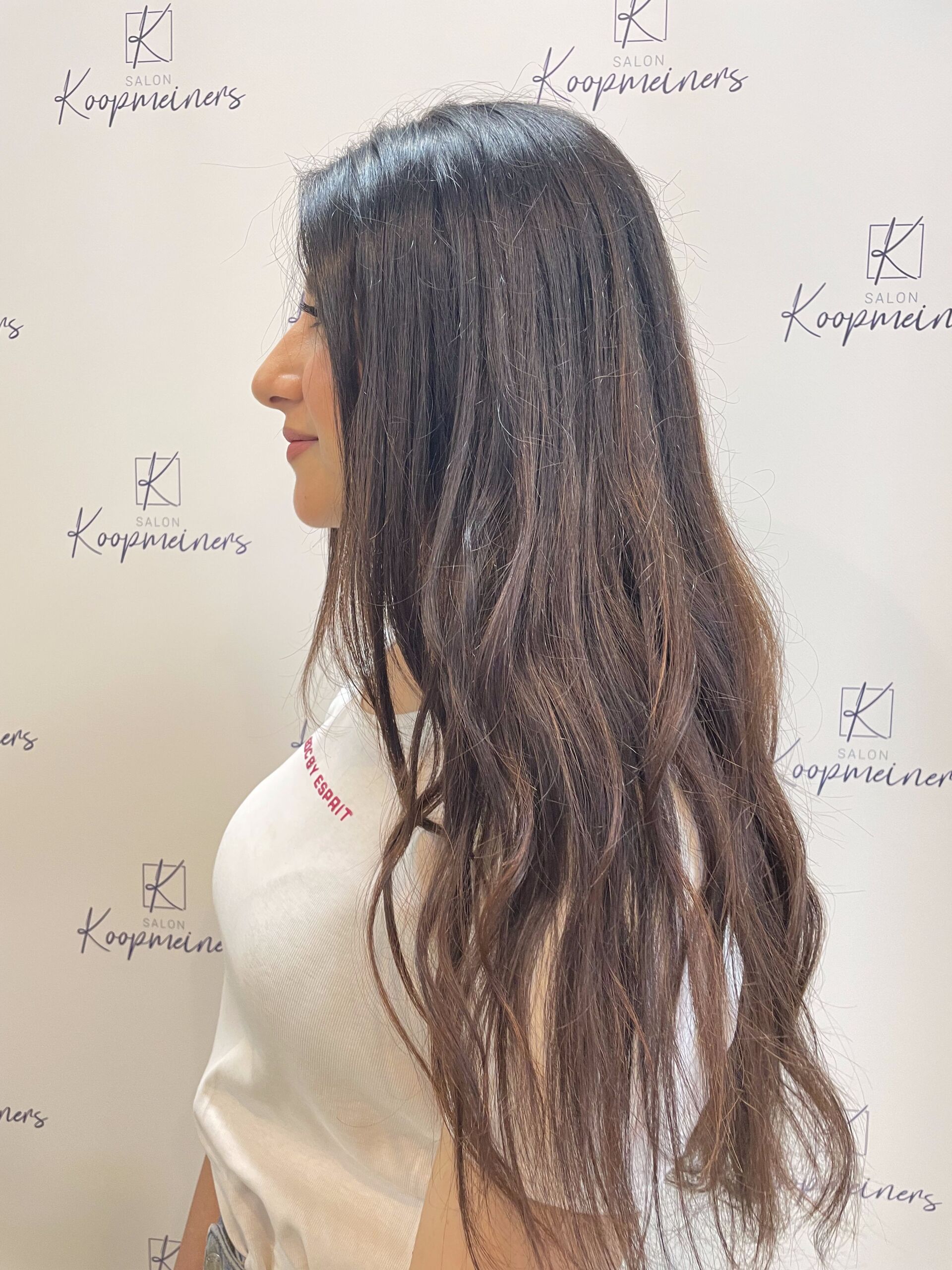 Frau mit langem, dunklem, welligem Haar steht im Profil vor einer weißen Wand mit Logos.