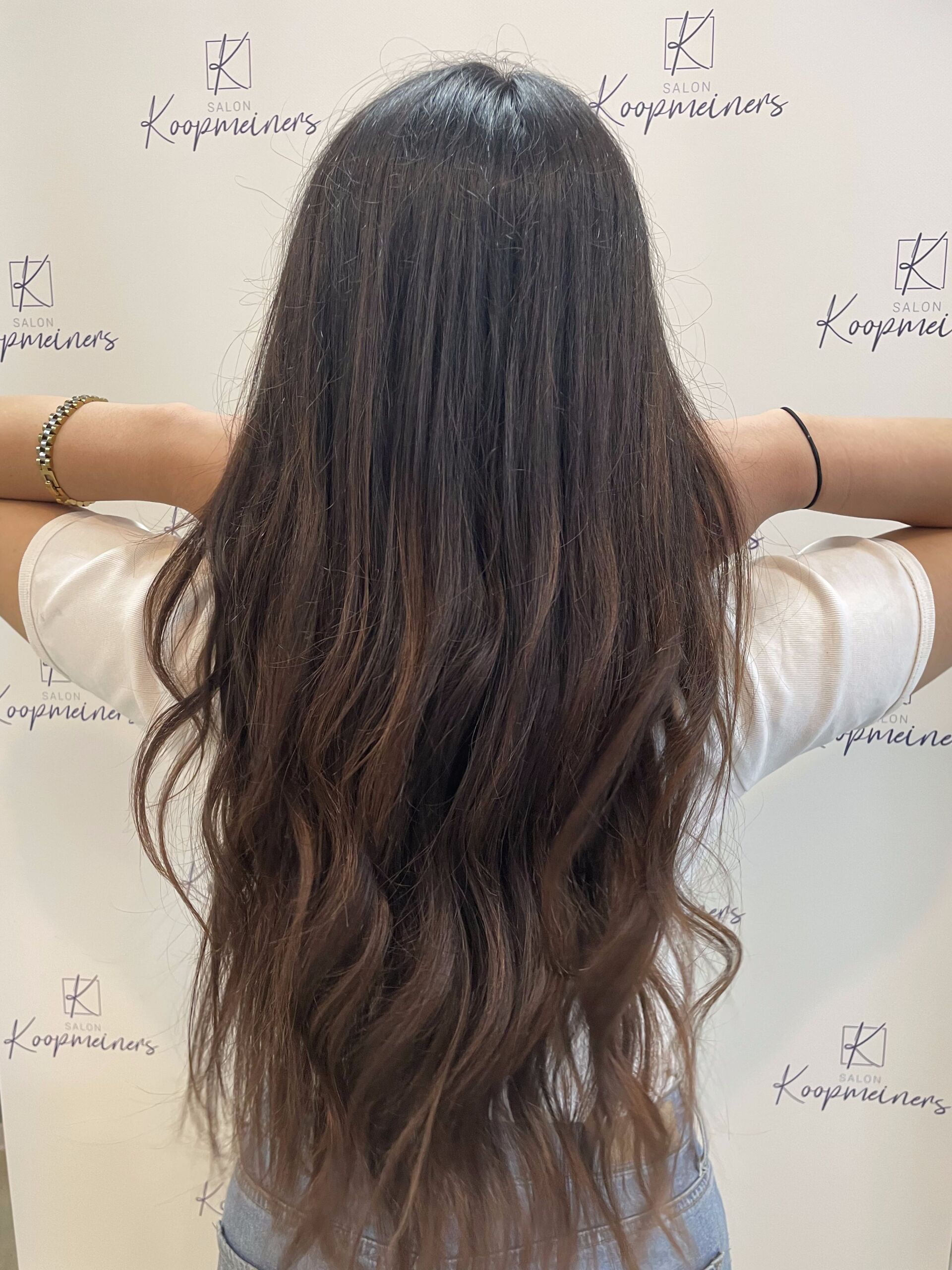 Frau mit langem, welligem braunem Ombré-Haar, die Arme hinter dem Rücken erhoben, vor einer weißen Wand.