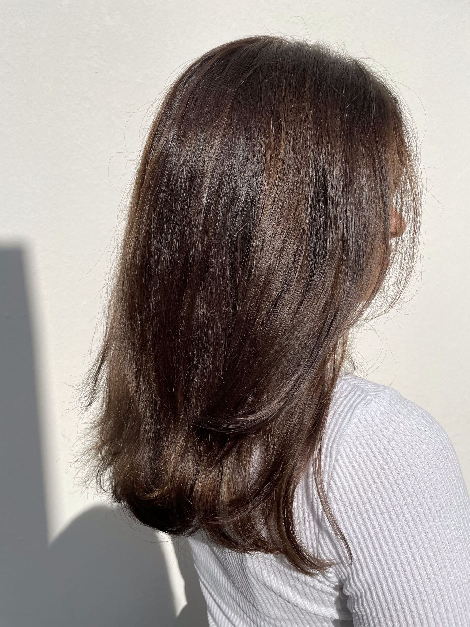 Person mit braunem Haar in einem grauen Pullover vor einer weißen Wand, beleuchtet vom Sonnenlicht.