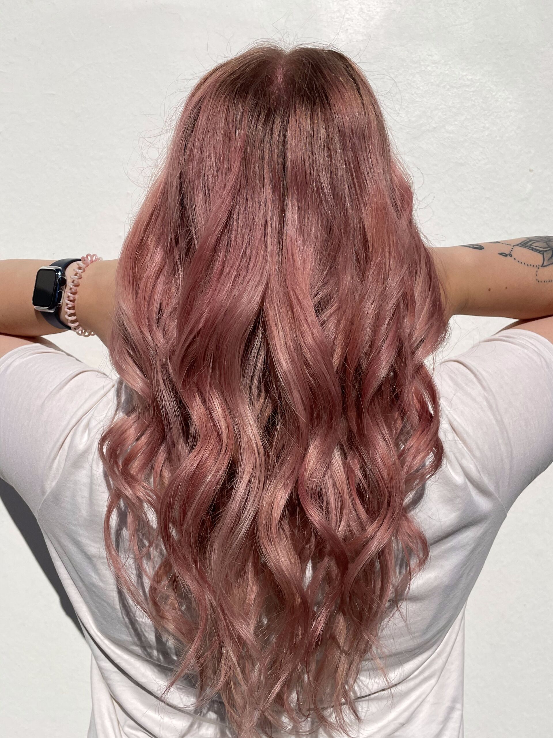 Frau mit langem, welligem, rosafarbenem Haar, die Arme hinter dem Kopf verschränkt, trägt ein weißes Hemd.