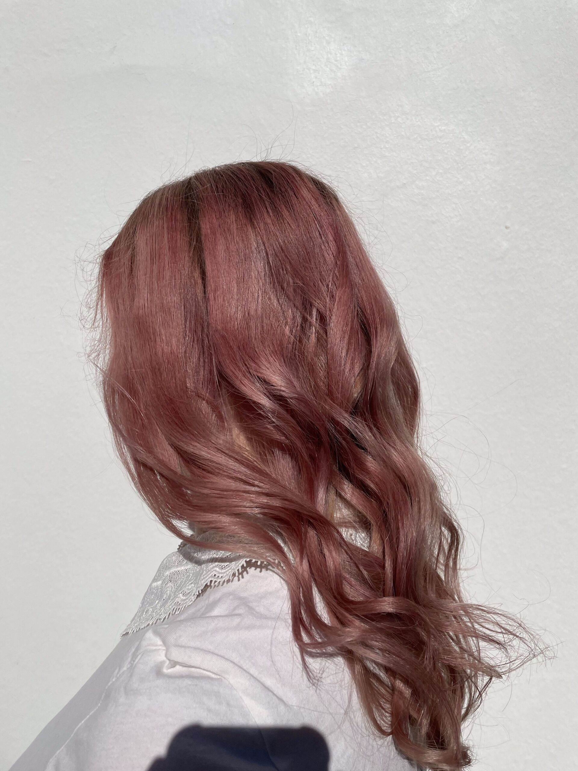 Person mit welligem, malvenfarbenem Haar, trägt ein weißes Hemd, von hinten vor einem weißen Hintergrund gesehen.