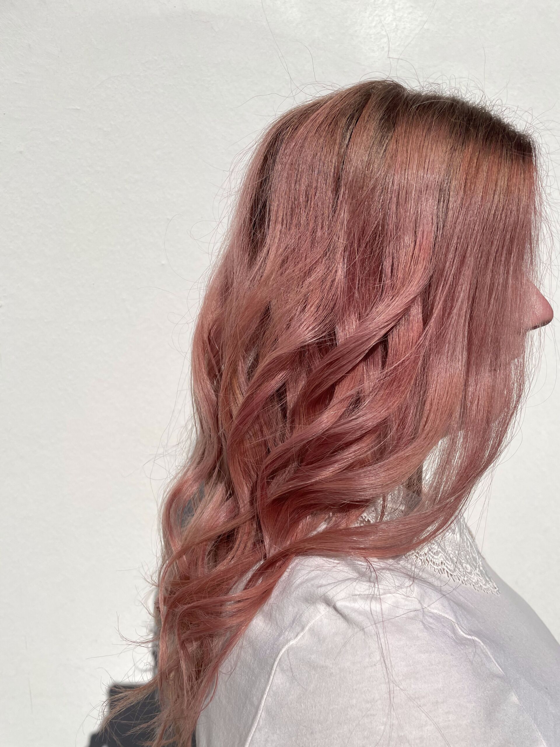 Frau mit welligem, rosa Haar und weißer Bluse, Profilansicht vor weißer Wand.