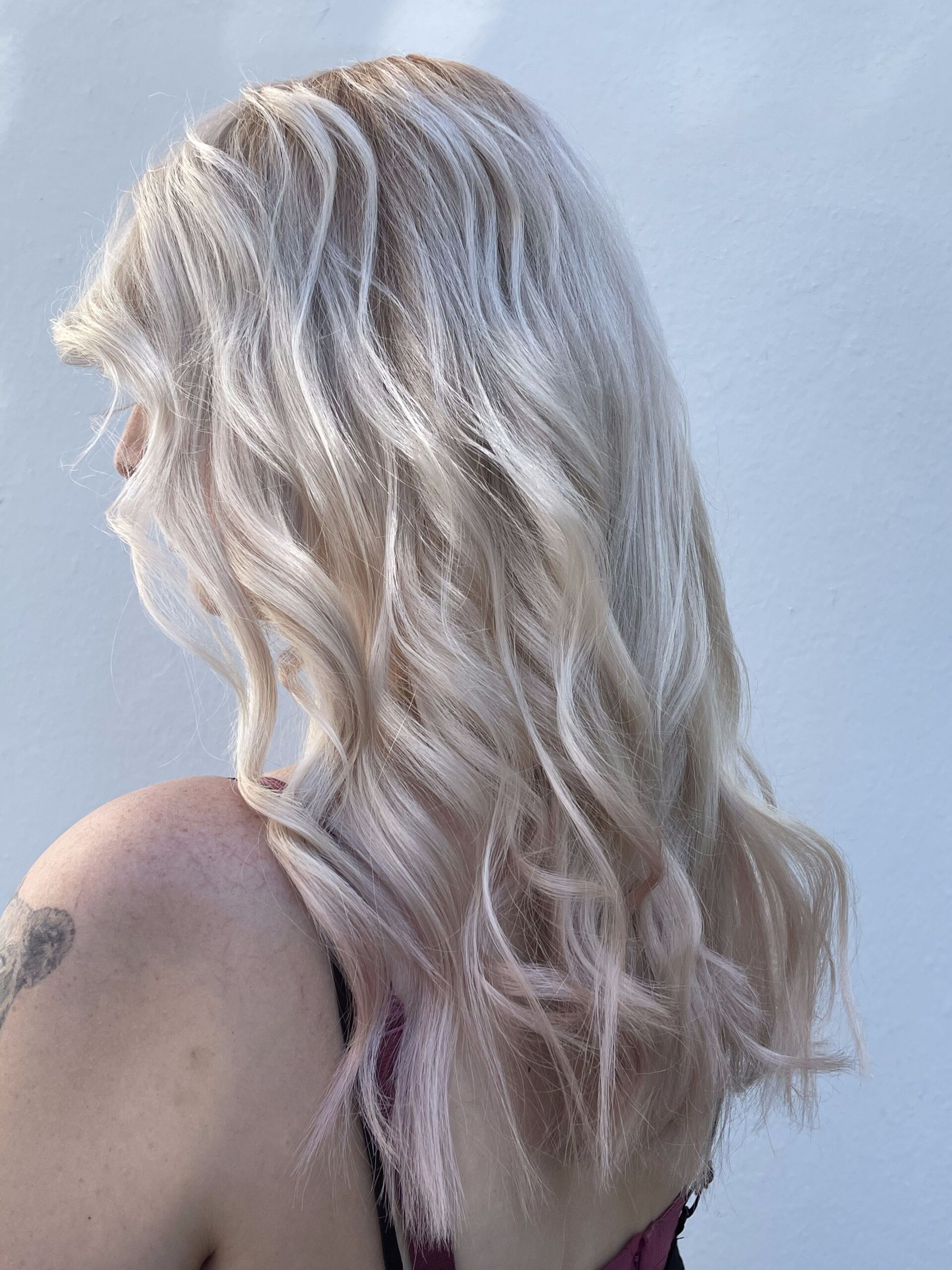 Frau mit blondem, welligem Haar, Schultertattoo und kastanienbraunem Oberteil.