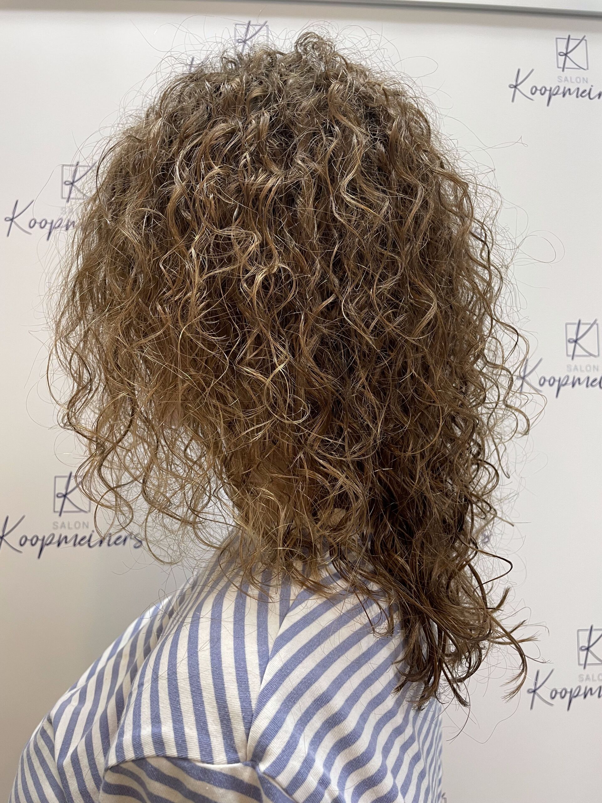 Person mit langem, lockigem, braunem Haar, die ein blau-weiß gestreiftes Hemd trägt und vor einer weißen Wand steht.