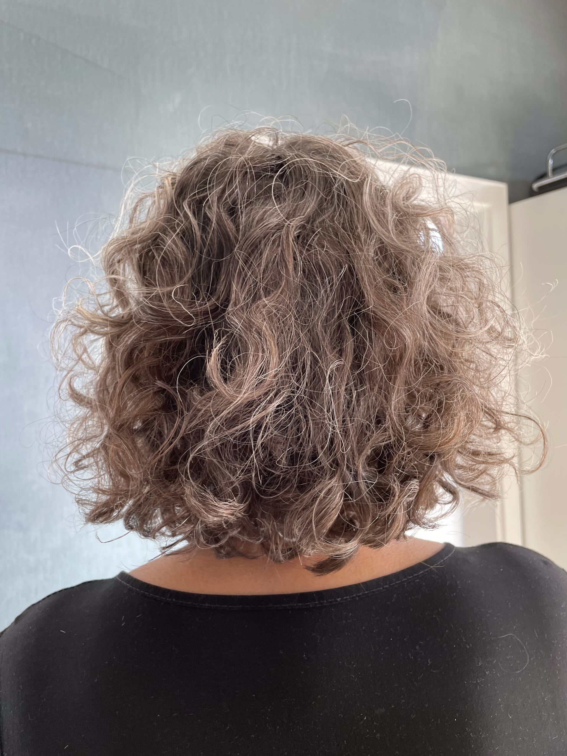 Person mit lockigem braunem Haar, bedeckt mit einer weißen Substanz, möglicherweise Schuppen, vor einer hellen Wand.