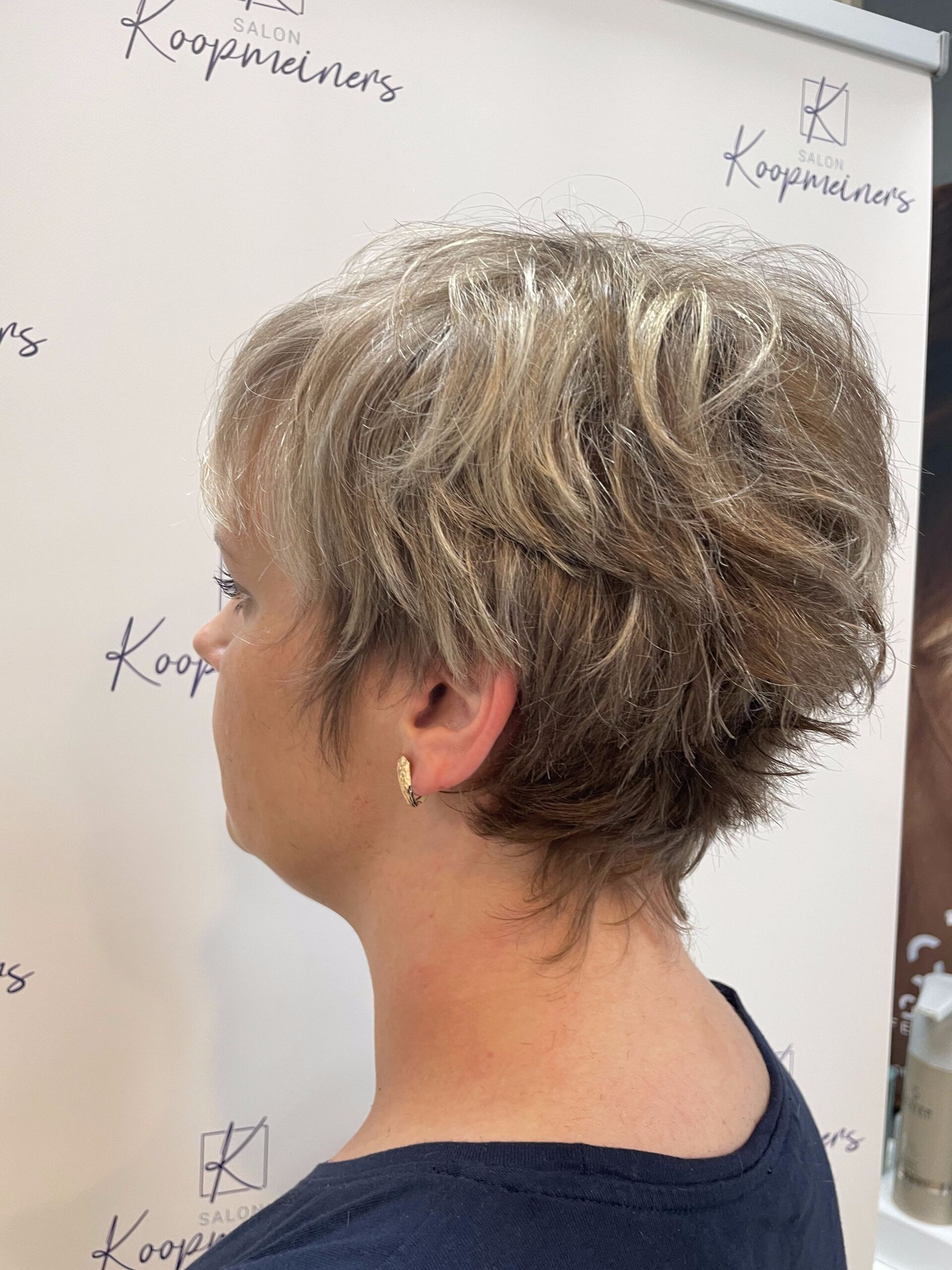 Frau mit kurzem, strukturiertem, gestuftem graublondem Haar; Profilansicht. Dunkelblaues Oberteil