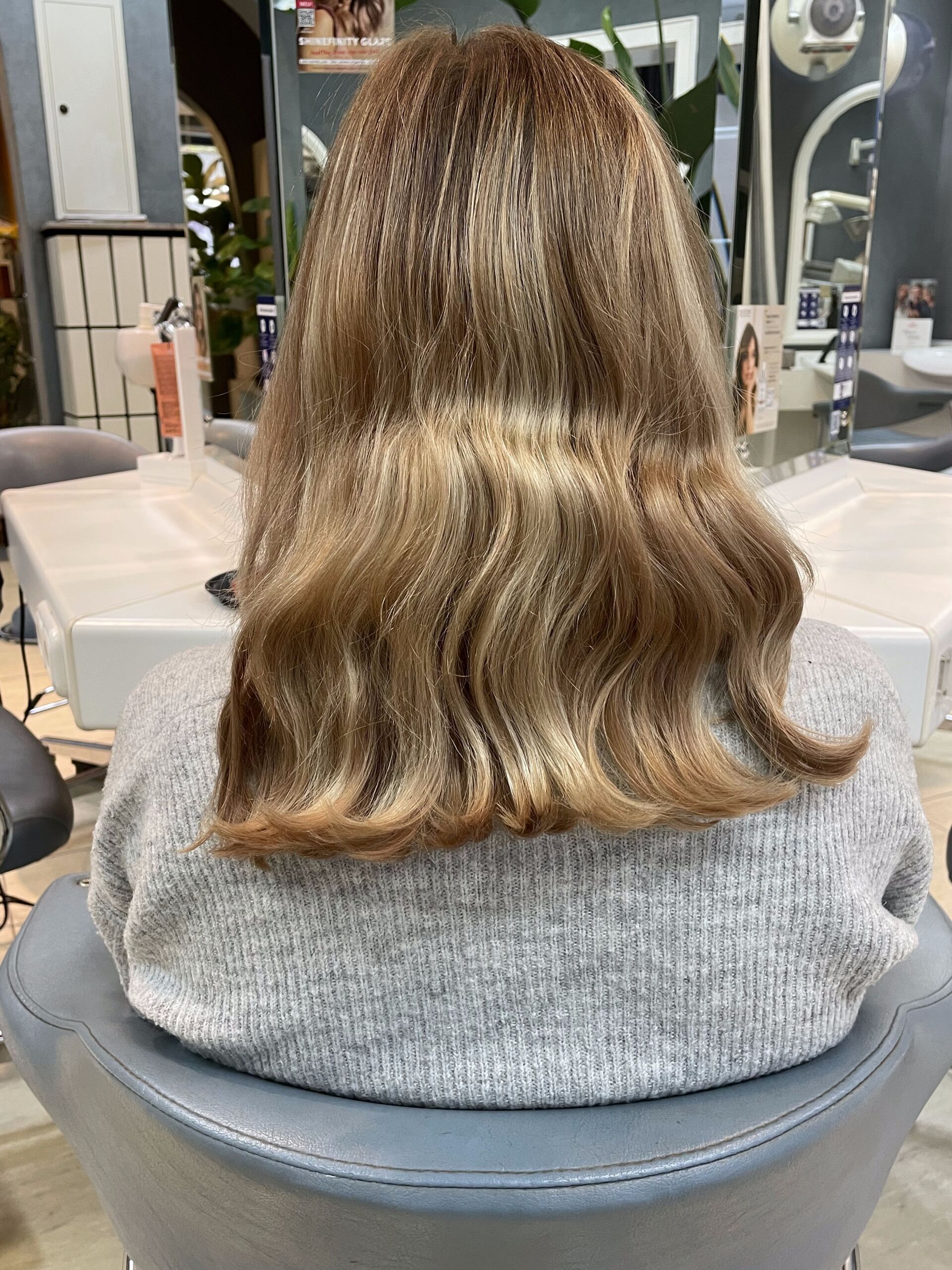 Eine Frau in einem grauen Pullover sitzt in einem Friseurstuhl, ihr hellbraunes, welliges Haar hat herabfallende Strähnchen.